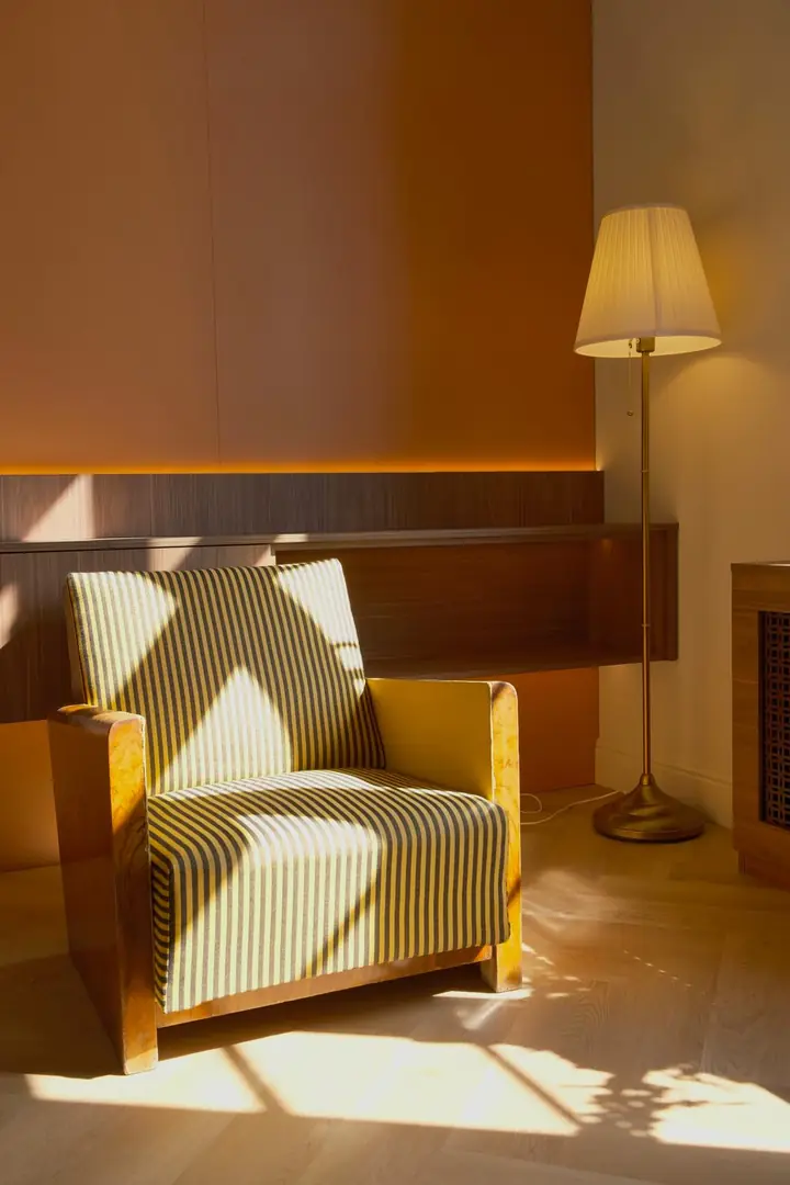 Un fotoliu Art Deco în dungi, în soarele de dimineață; umbra cerceveii cade pe spătar și pe parchet. Lângă el, un lampadar de alamă aprins.
