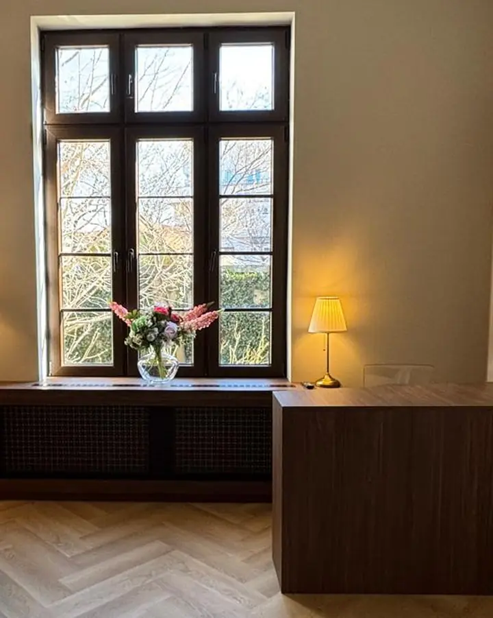 Fereastra înaltă cu cercevele de lemn, încadrată de două lămpi aprinse, cu un buchet de flori pe calorifer.