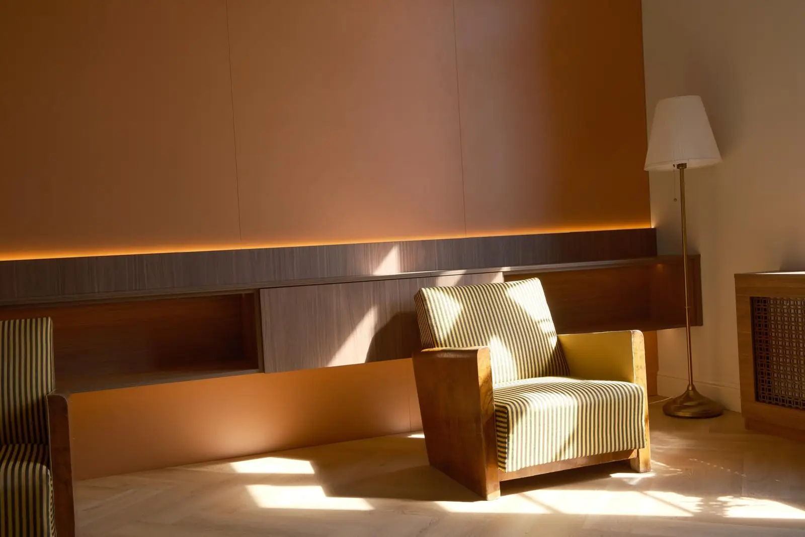 Sala de așteptare dimineața: un fotoliu Art Deco tapițat în dungi, în lumina care intră printre cercevele, lângă un lampadar de alamă. În spate, peretele ocru, cu o bandă de lumină caldă ascunsă sub raftul de nuc.